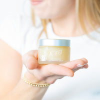 A person holding a small jar of Deep Moisture Cleansing Balm with their fingers lightly touching the sides.