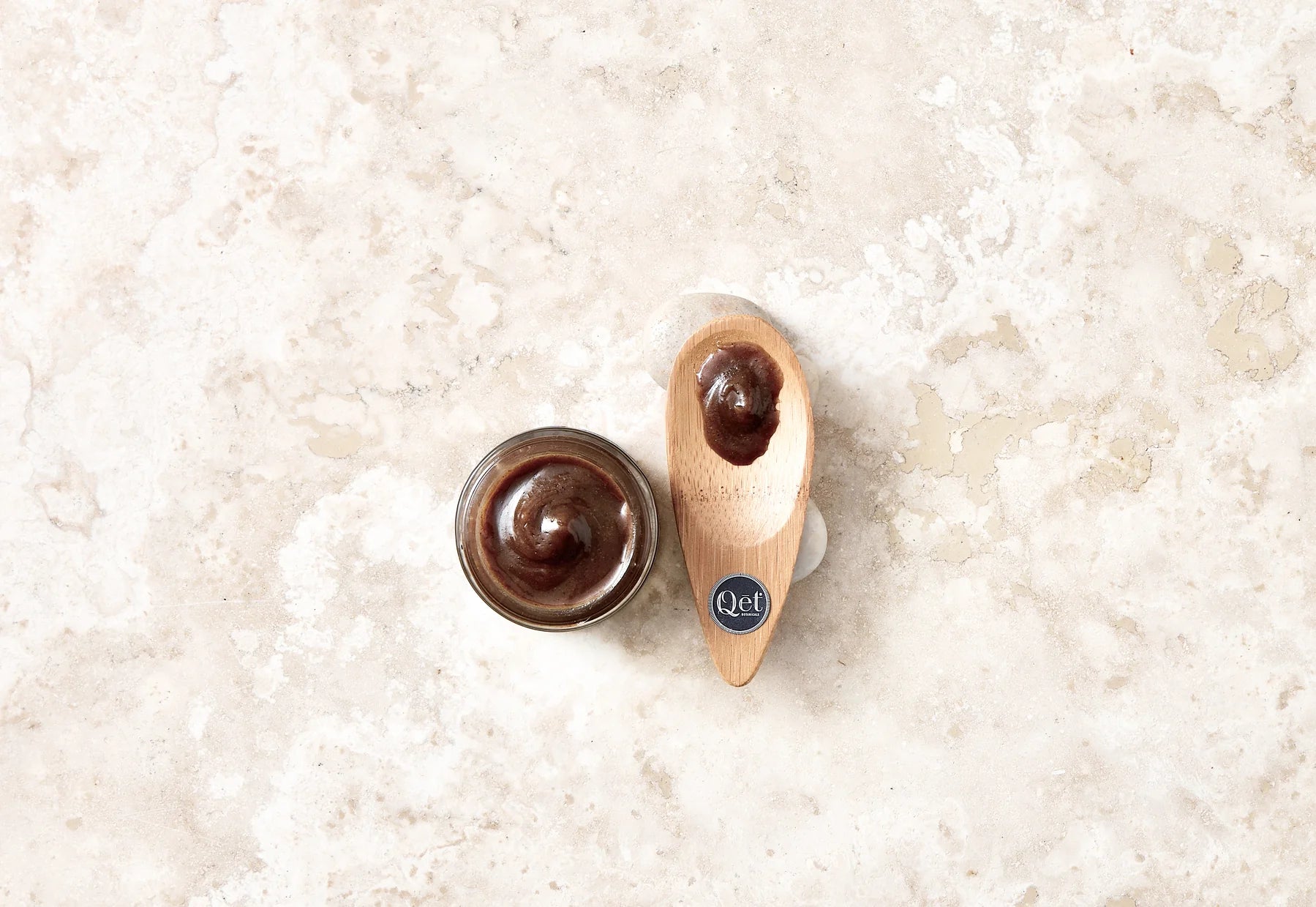 A jar and a wooden spoon rest on a marble surface, both topped with a swirl of chocolate spread. The spoon features a small, dark blue sticker.