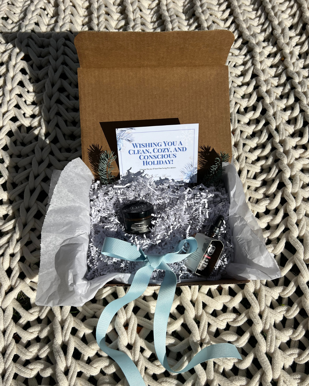 Open cardboard box on knitted surface, containing shredded paper, small evergreen sprigs, candles, soft blue ribbon, and a holiday card.