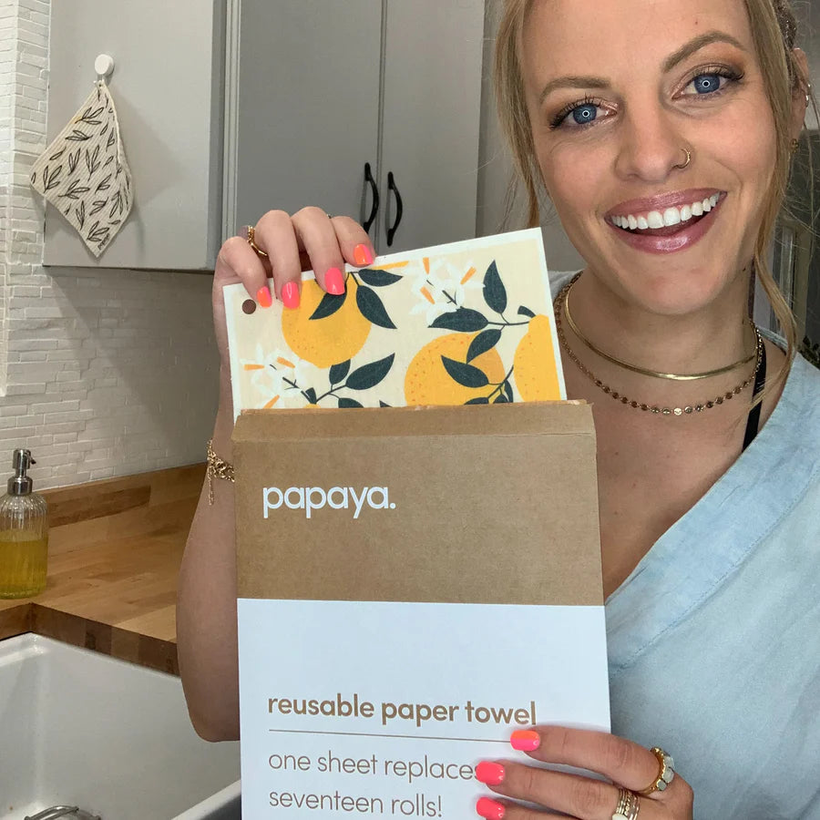 Smiling woman holds a package of "papaya" reusable paper towels with a design of lemons and leaves. Kitchen setting with a cozy, eco-friendly vibe.