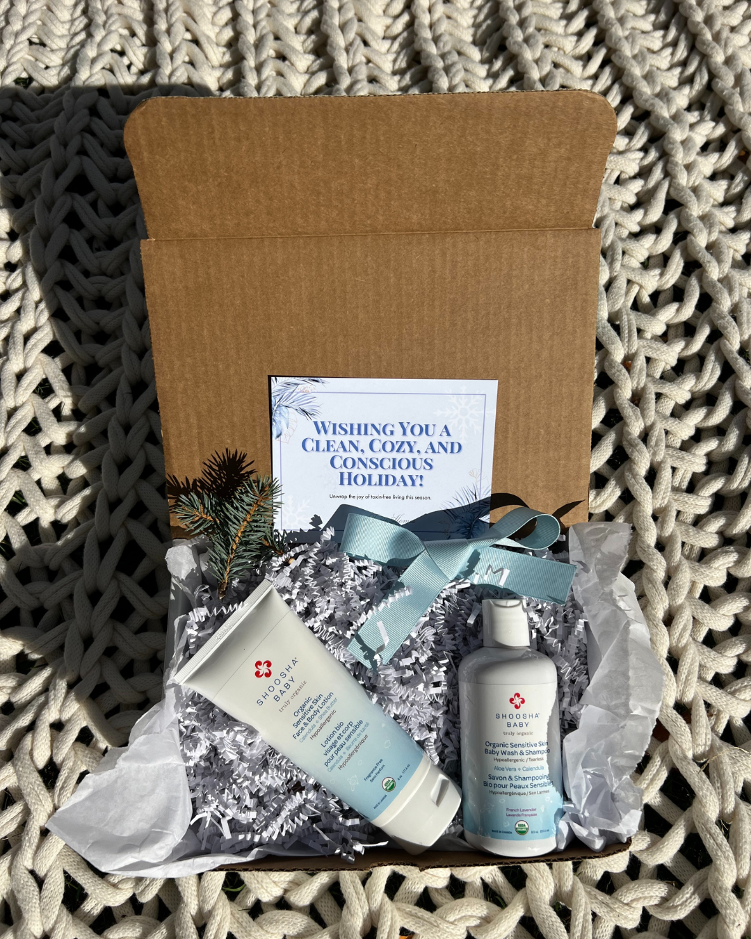 Gift box with skincare products on white crinkle paper, including a bottle and tube. A card reads "Wishing You a Clean, Cozy, and Conscious Holiday."