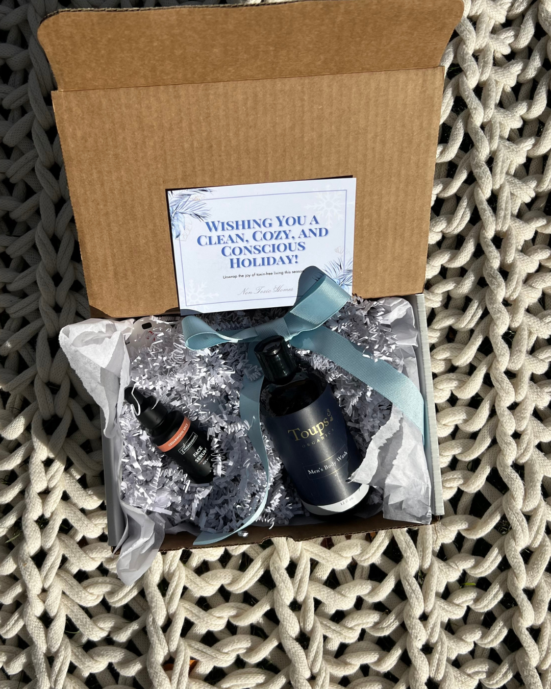 Open cardboard box on a knitted surface contains a bottle, a small dropper, and a card with “Wishing You a Clean, Cozy, and Conscious Holiday!” in snowy font.