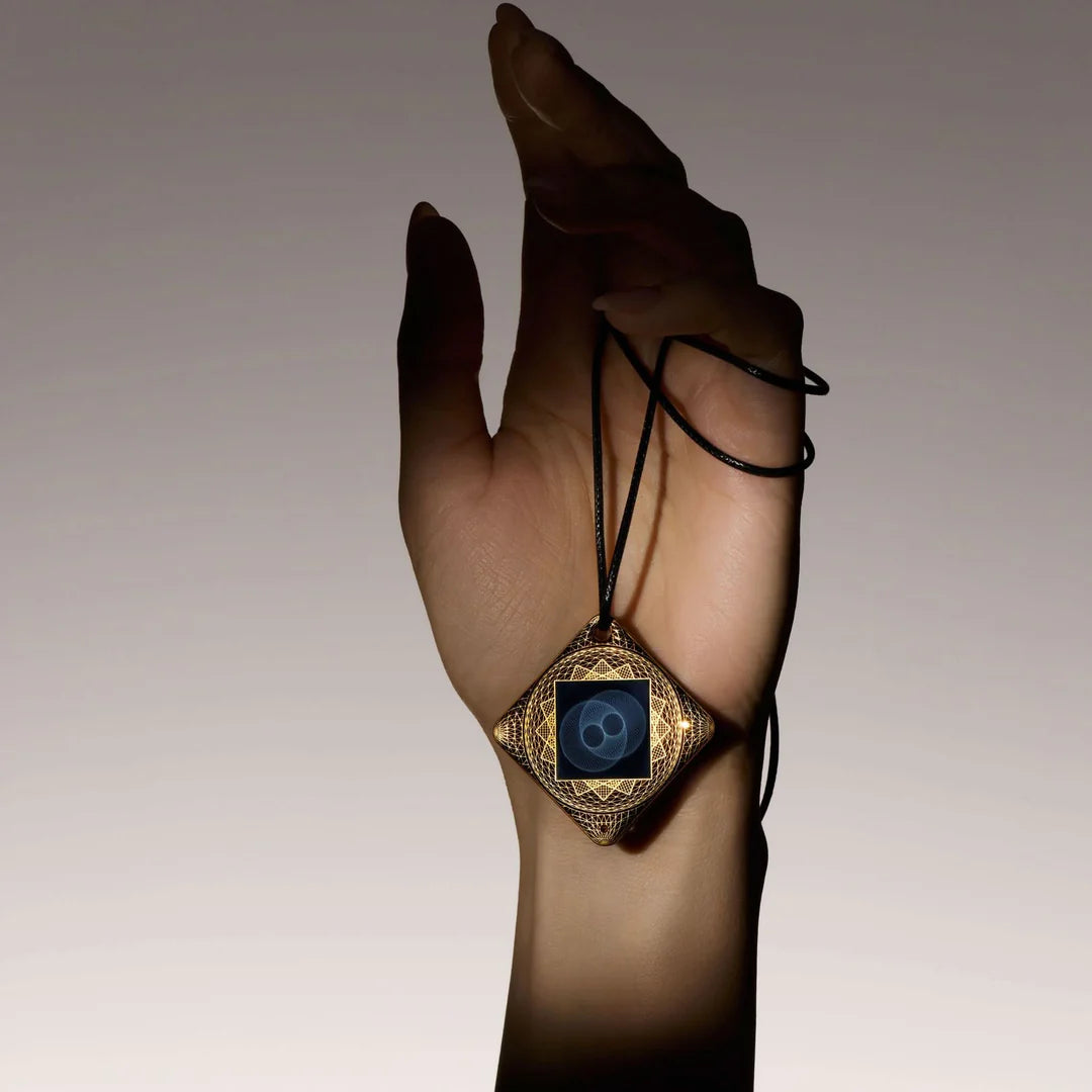 A hand gently holds an ornate, square pendant with intricate gold designs and black cord. The image conveys elegance and mystery against a soft gray background.