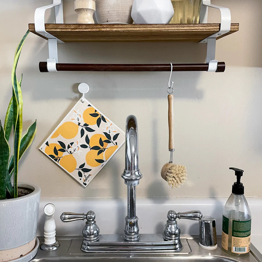 A cozy kitchen sink setup features a silver faucet, soap bottle, and succulent plant. Above, a wooden shelf holds decor, hanging a yellow-patterned dish towel and brush.