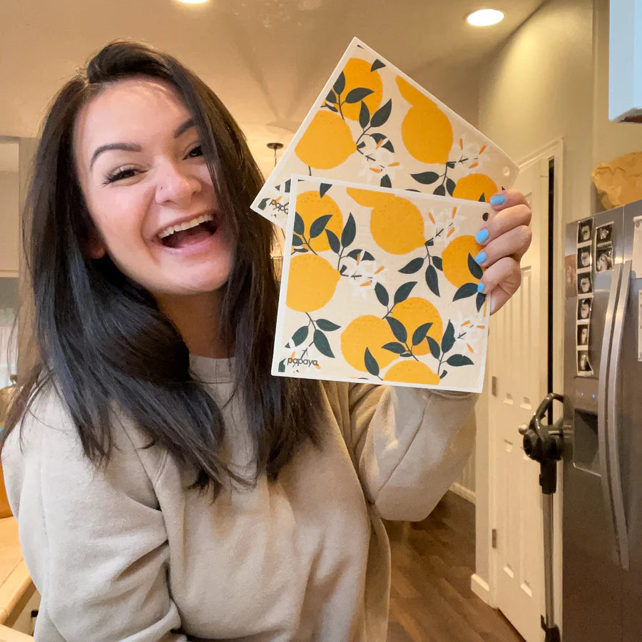 A woman smiling broadly holds three art prints featuring a bright yellow lemon pattern with green leaves. She stands in a cozy kitchen, exuding joy.