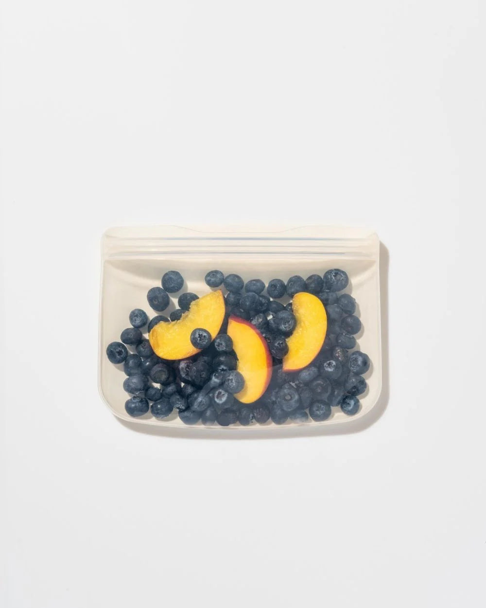 A reusable, clear silicone bag filled with fresh blueberries and peach slices on a white background. The image conveys freshness and eco-friendliness.
