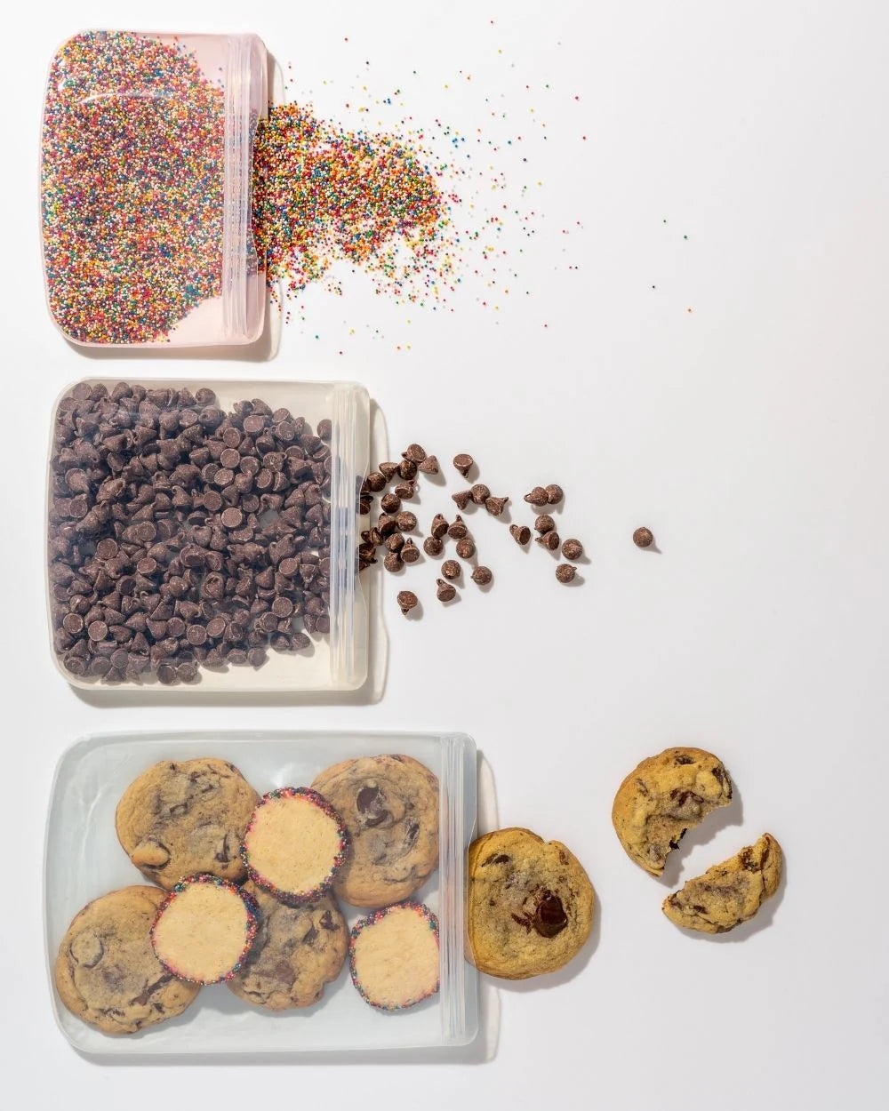 Three open jars are arranged vertically on a white surface. The top has colorful sprinkles spilling out, the middle shows chocolate chips scattered, and the bottom contains cookies, including a few broken pieces beside the jar.