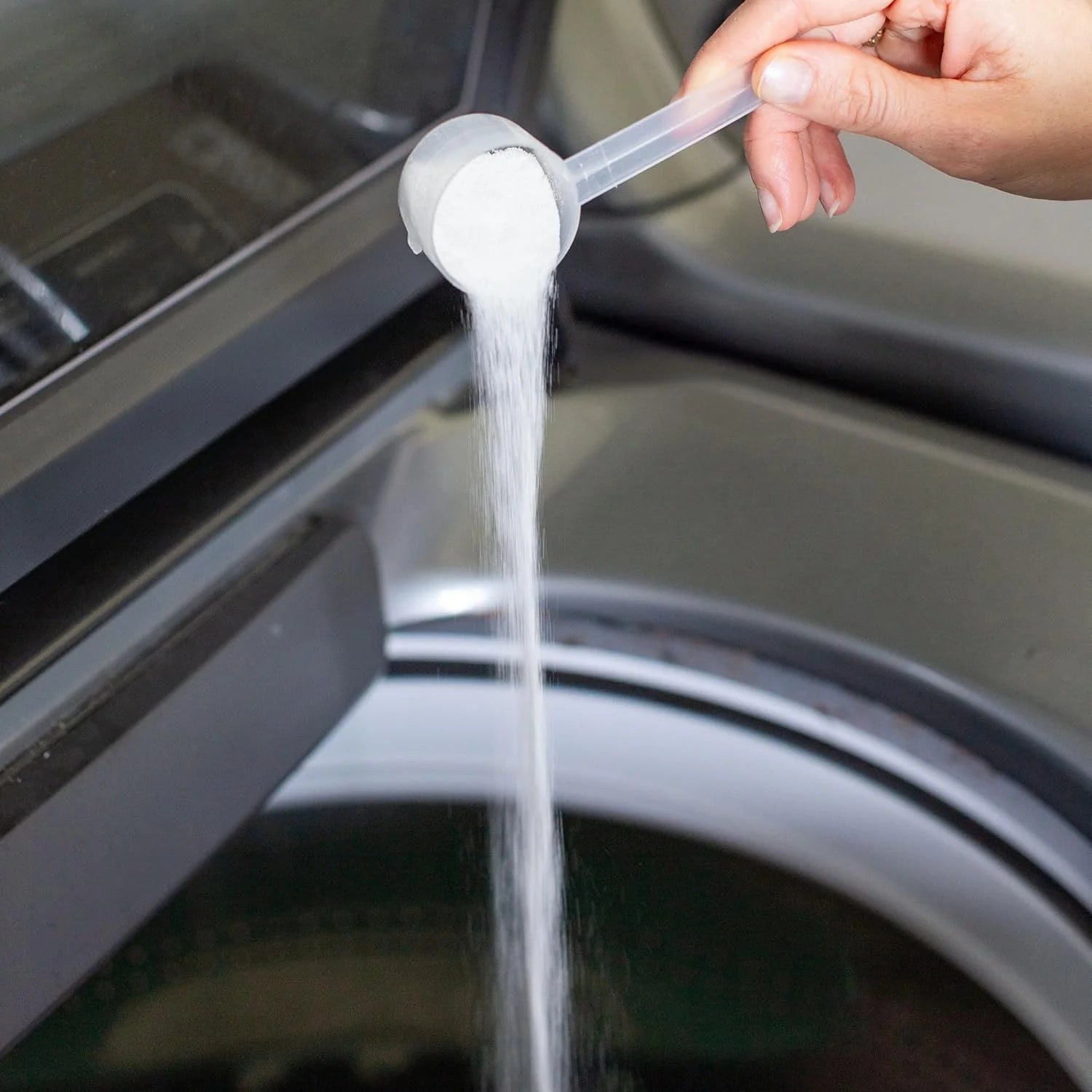 A hand pours laundry detergent powder from a scoop into a washing machine. The scene suggests household chores and cleanliness.