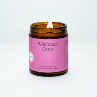 A photo of a lit Wildflower Citrus scented candle with a pink label, made by Fontana Candle Co. in a brown jar.