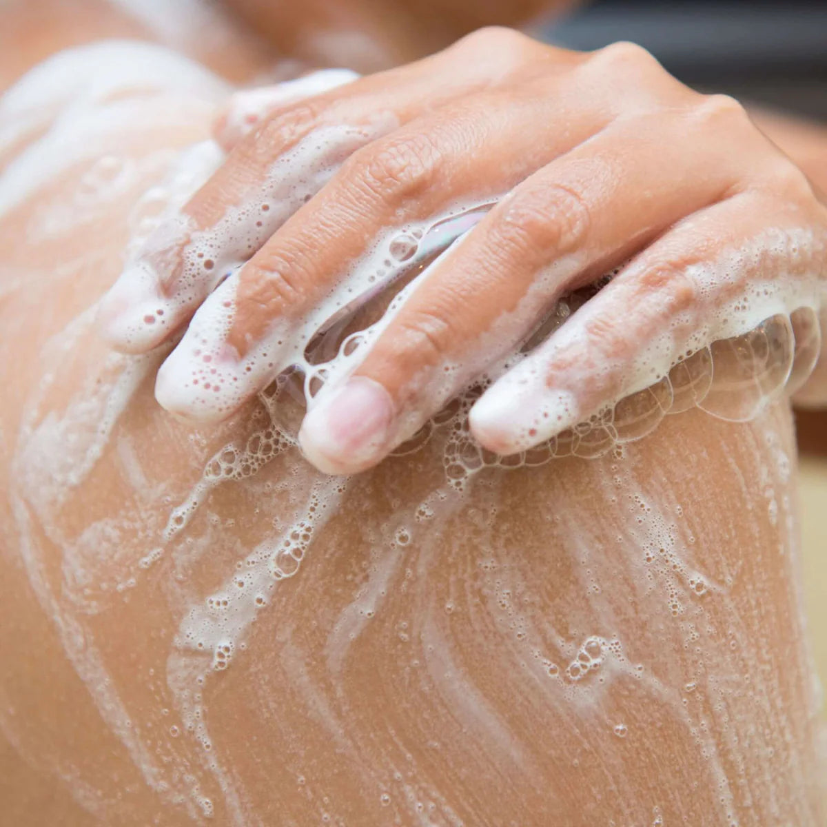 A hand gently scrubs a shoulder with soap, creating white suds and bubbles. The image conveys a sense of cleanliness and self-care.