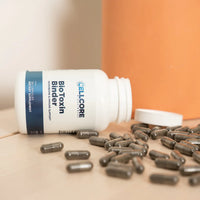 A bottle of 'BioToxin Binder' supplement with capsules spilling out in front of it, on a wooden surface with a neutral background.