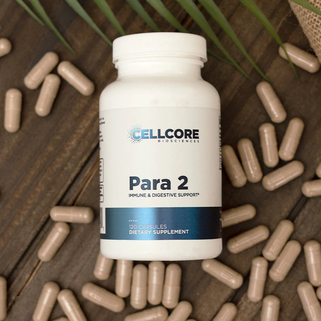 A bottle of 'Para 2' dietary supplement by Cellcore Biotech, surrounded by capsules on a wooden surface with a green leafy herb in the background.