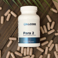 A bottle of 'Para 2' dietary supplement by Cellcore Biotech, surrounded by capsules on a wooden surface with a green leafy herb in the background.