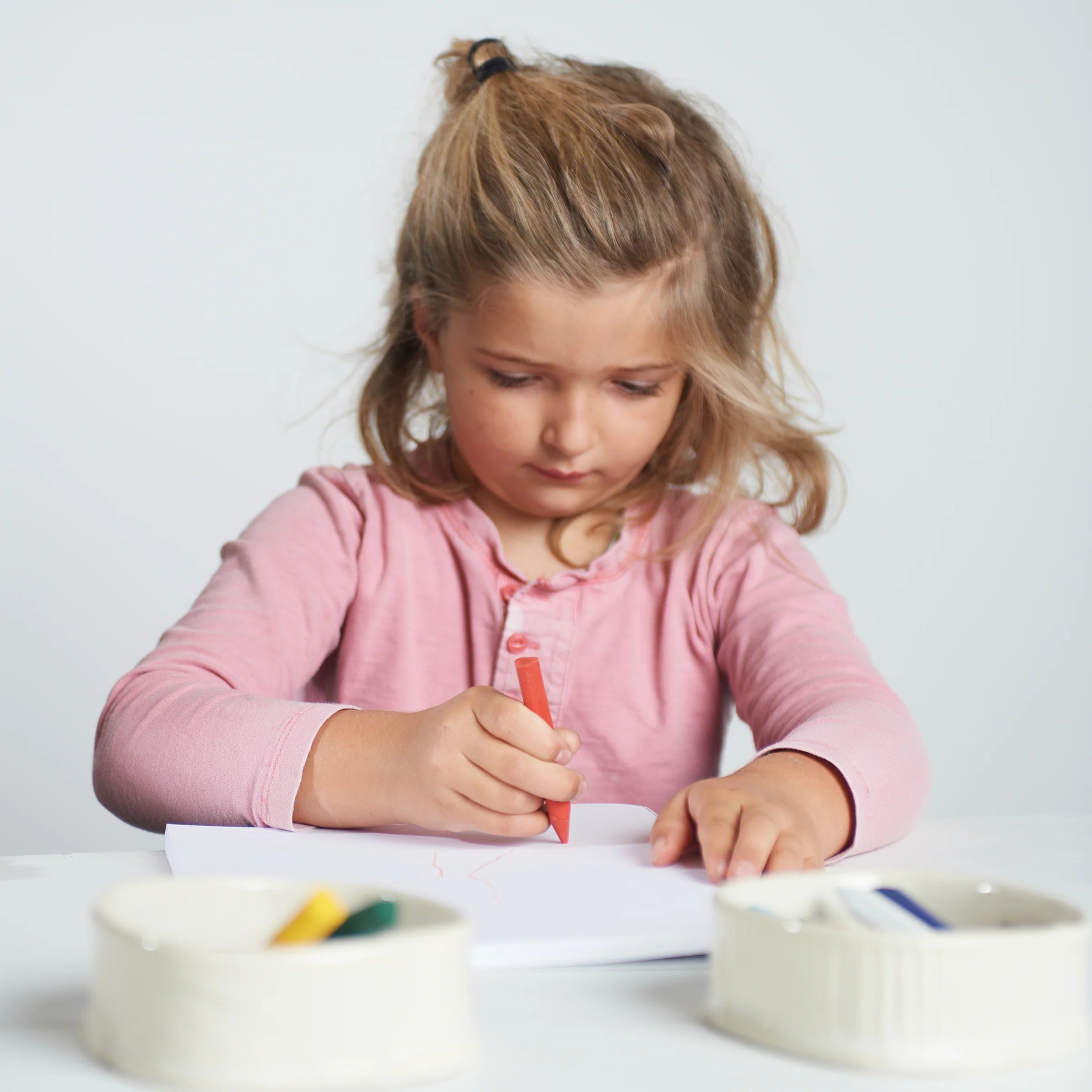 Young girl in a pink shirt writing on paper with a red crayon.