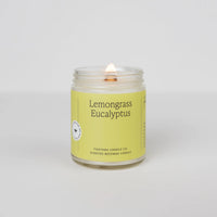 A clear glass candle with a label reading 'LEMONGRASS EUCALYPTUS', with a lit wick and a small flame, placed on a neutral background.