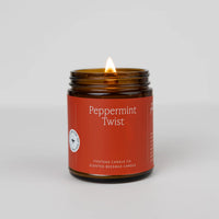 A peppermint twist essential oil candle with a lit flame, housed in a brown glass jar with a label that includes the product name and brand.