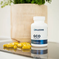 A bottle of GCO supplements with yellow capsules spilling out in front of it, placed on a reflective surface with a plant in the background.