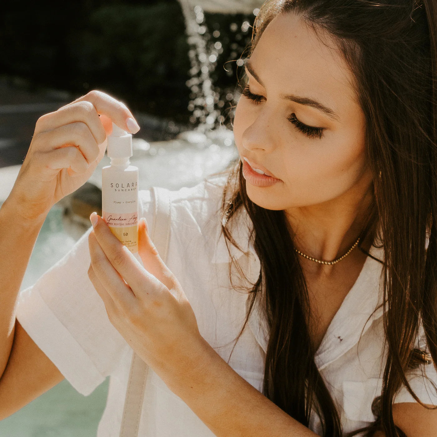 A woman examines a small skincare bottle outdoors, dressed in a white shirt. Sunlight and a blurred fountain in the background create a serene, focused mood.