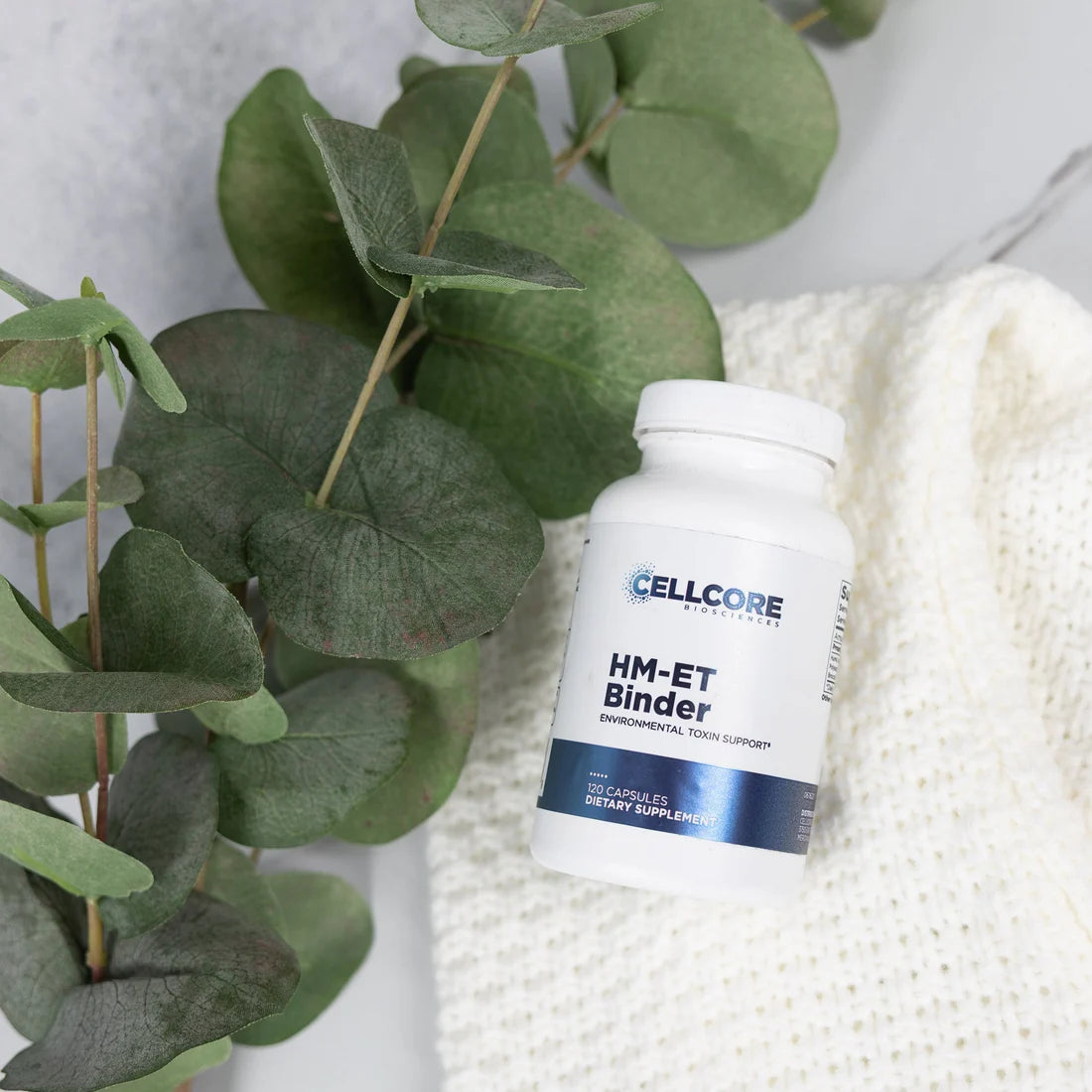 A bottle of HM-ET Binder supplement from CELLCORE, placed on a white fabric with a green, leafy plant in the background.