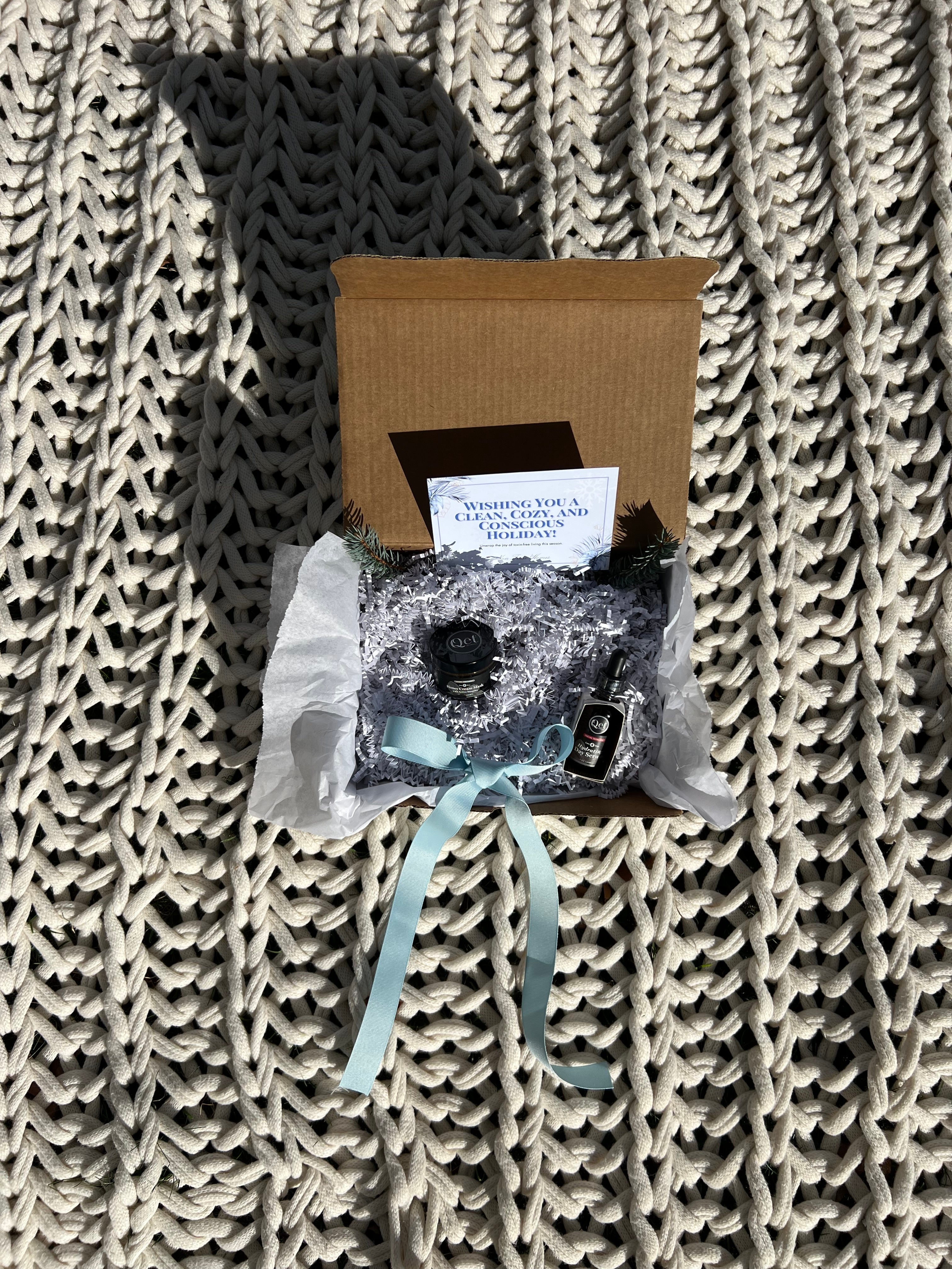 Open cardboard gift box on a textured knit blanket. Inside, a card, small black jar, and a bottle are nestled in shredded paper with a blue ribbon.