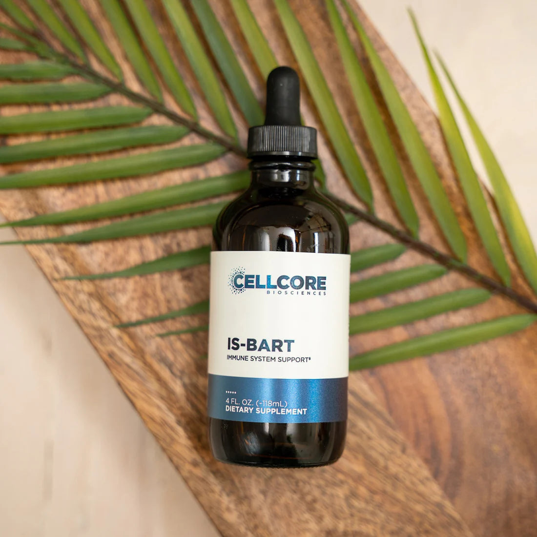A bottle of IS-BART tincture by CELLCORE, placed on a wooden surface with a green leaf in the background, indicating it is a dietary supplement.