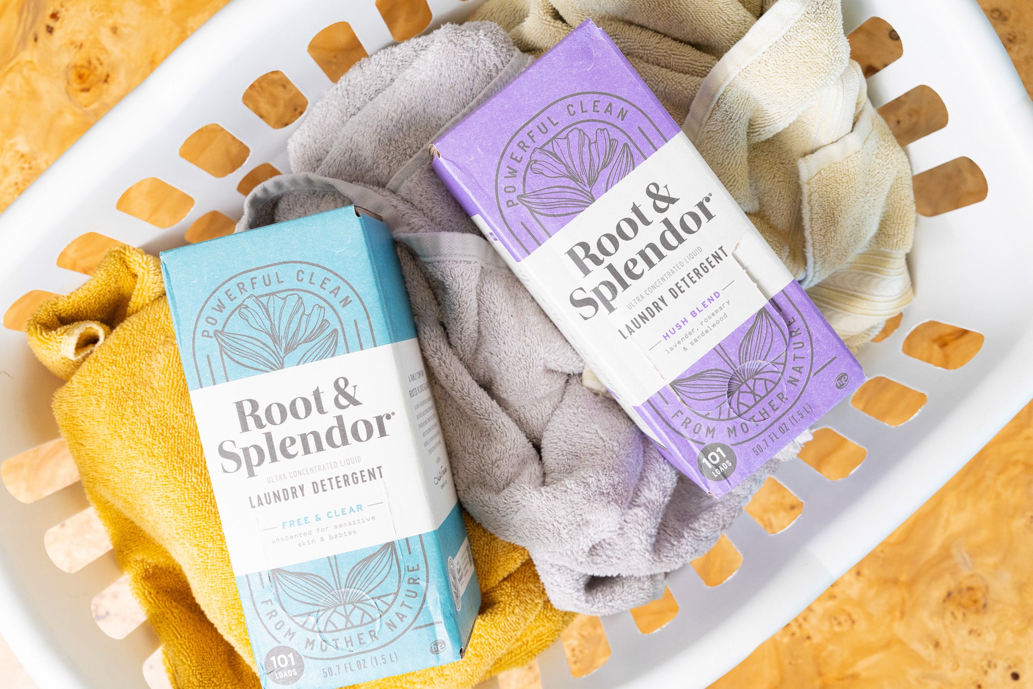 Two packages of Root and Splendor laundry detergent in a basket with laundry clothes. One package is labeled 'Hush Blend' with purple and gray design, and the other is 'Fresh & Clear' with a yellow and green design.