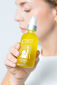 A person holding a yellow bottle of Activist Botanical Cleansing Oil with a pump dispenser.