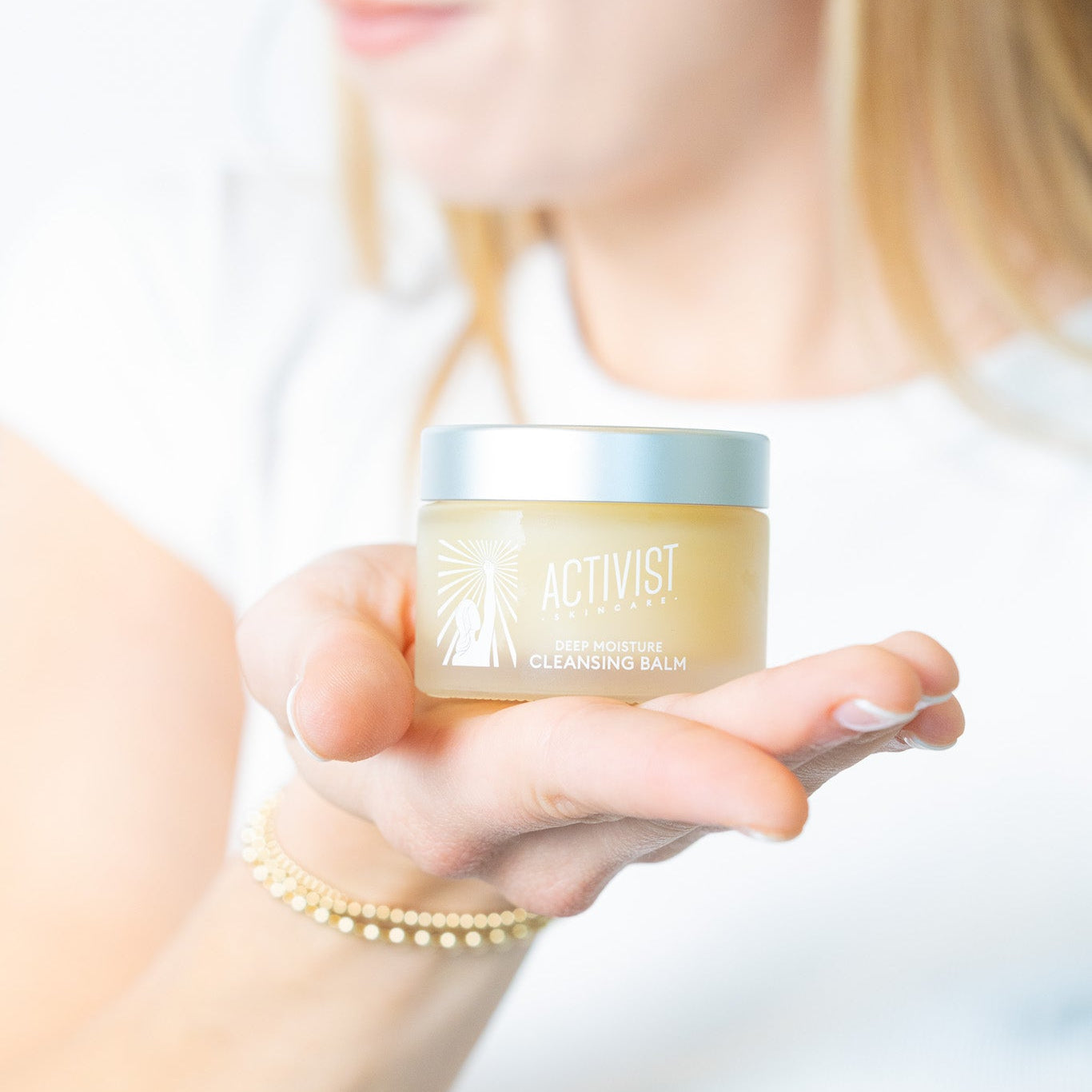 A person holding a small jar of Deep Moisture Cleansing Balm with their fingers lightly touching the sides.