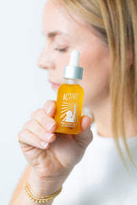 A person holding a bottle of face oil labeled 'Precious Oils Active Infusion' with a dropper, in front of a white background.