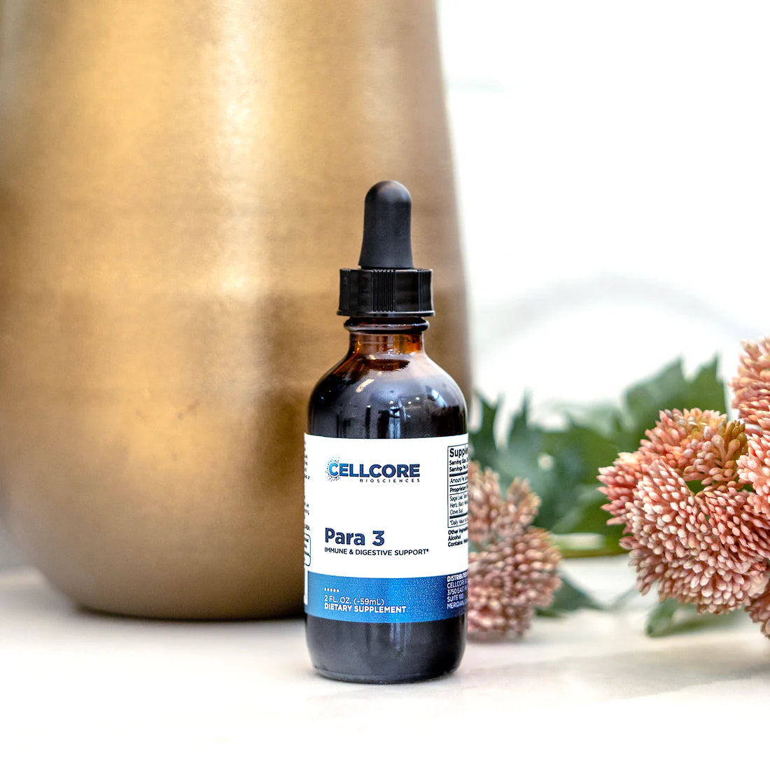 A bottle of 'Para 3' supplement with a dropper, placed on a white surface, with a metallic container and flowers in the background.