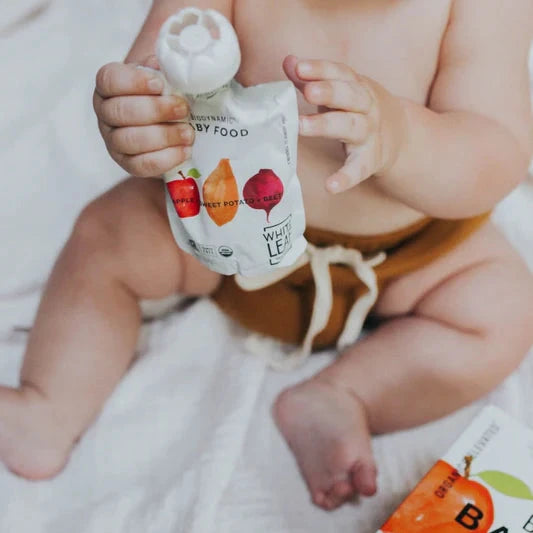 A baby sitting on a white blanket holds a pouch of organic baby food labeled with apple and carrot illustrations. The scene is calm and playful.