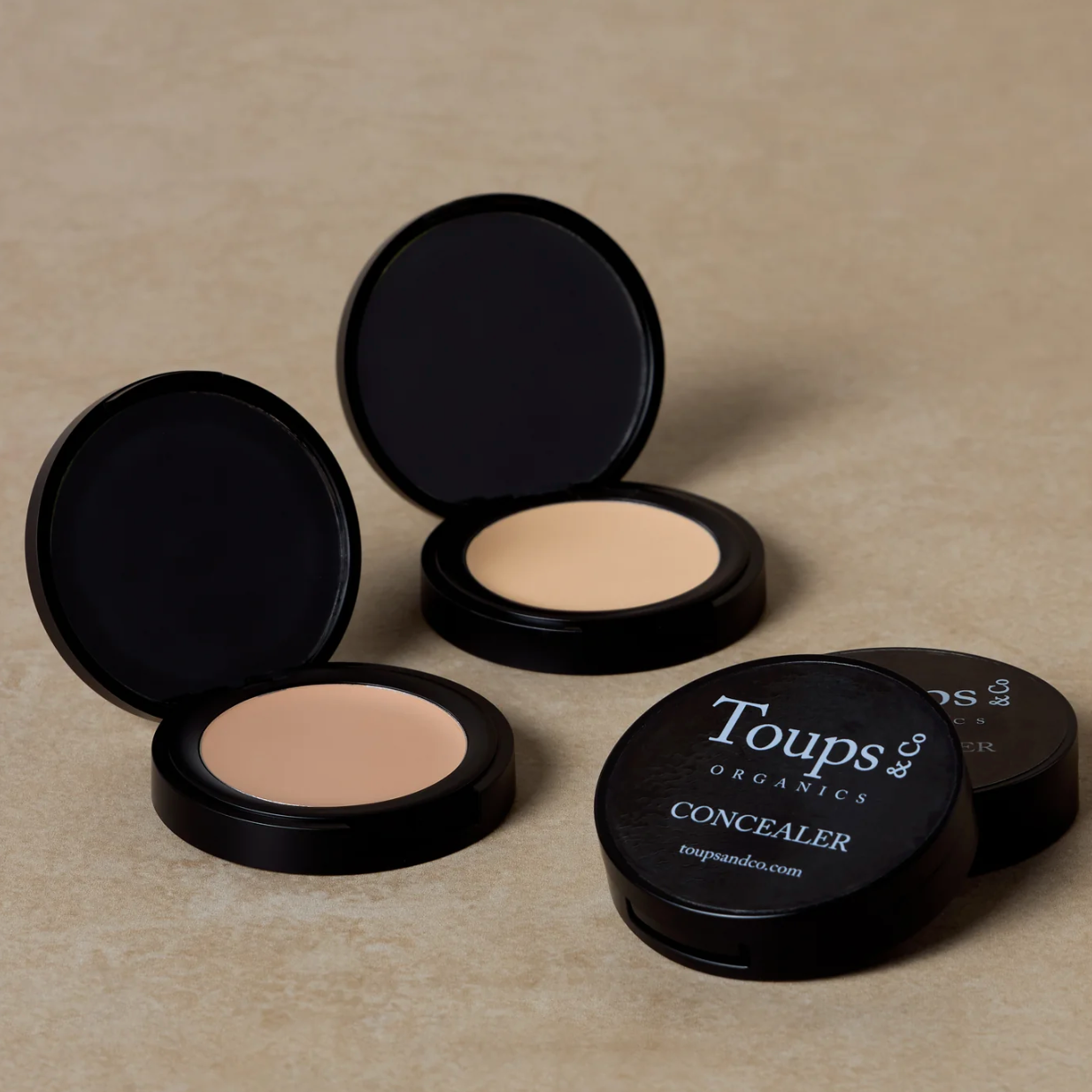 Two compact concealers with black lids, one open showing the product and the other closed, with the brand name 'Toups' visible on the packaging.