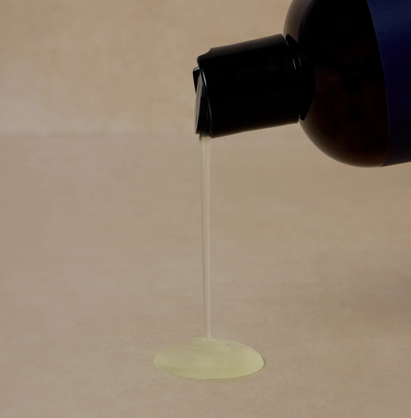 A dark bottle tipped, pouring a smooth, translucent liquid onto a beige surface. The image conveys simplicity and focus on texture.