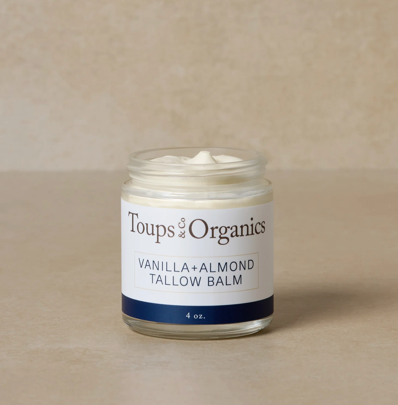 A glass jar containing Toups Organics Vanilla + Almond Tallow Balm, with a label indicating the product name and size (4 oz.).