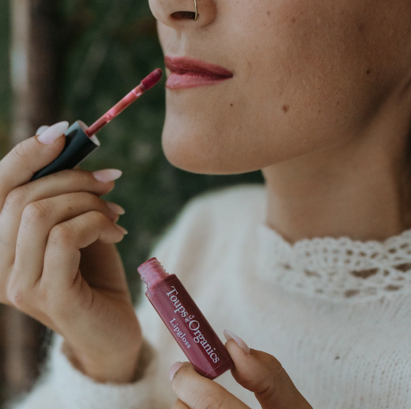 A close-up image of a person's face, specifically focusing on the lips, with a swatch of color indicating the organic lip gloss being applied.