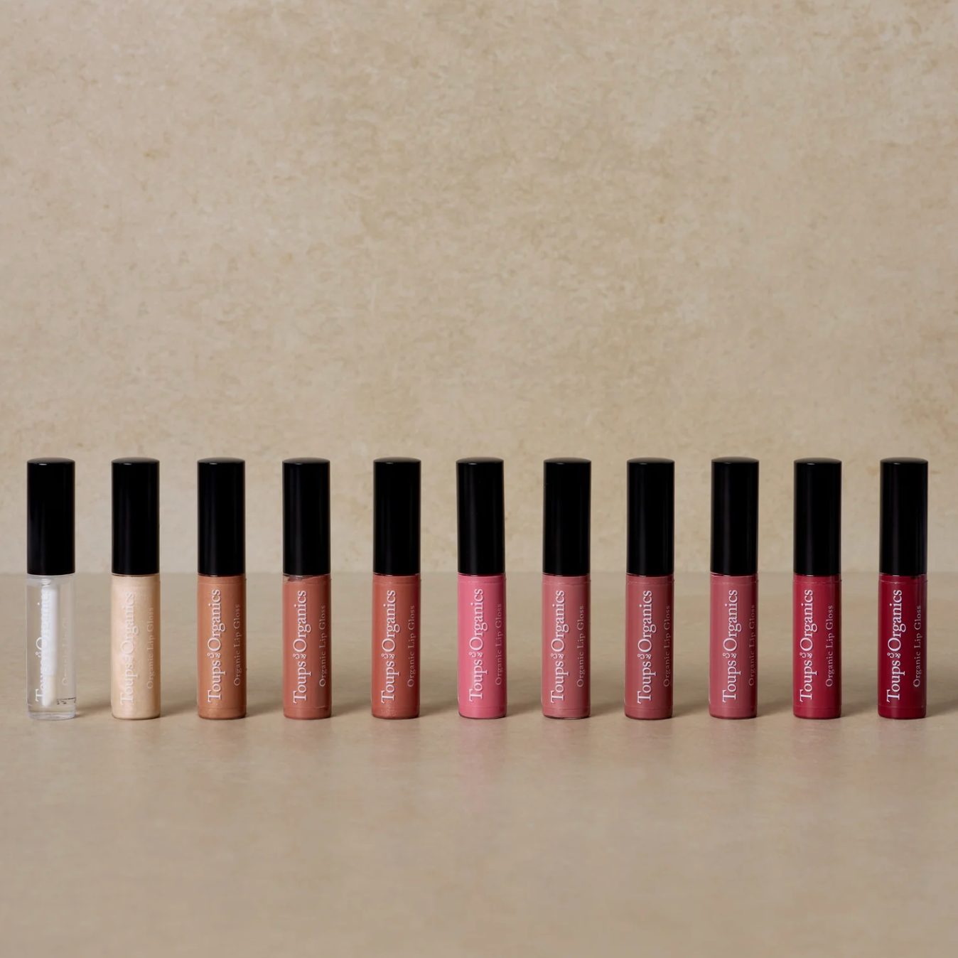 A collection of organic lip glosses in various shades, displayed in a row with the applicators in black color, on a beige background.