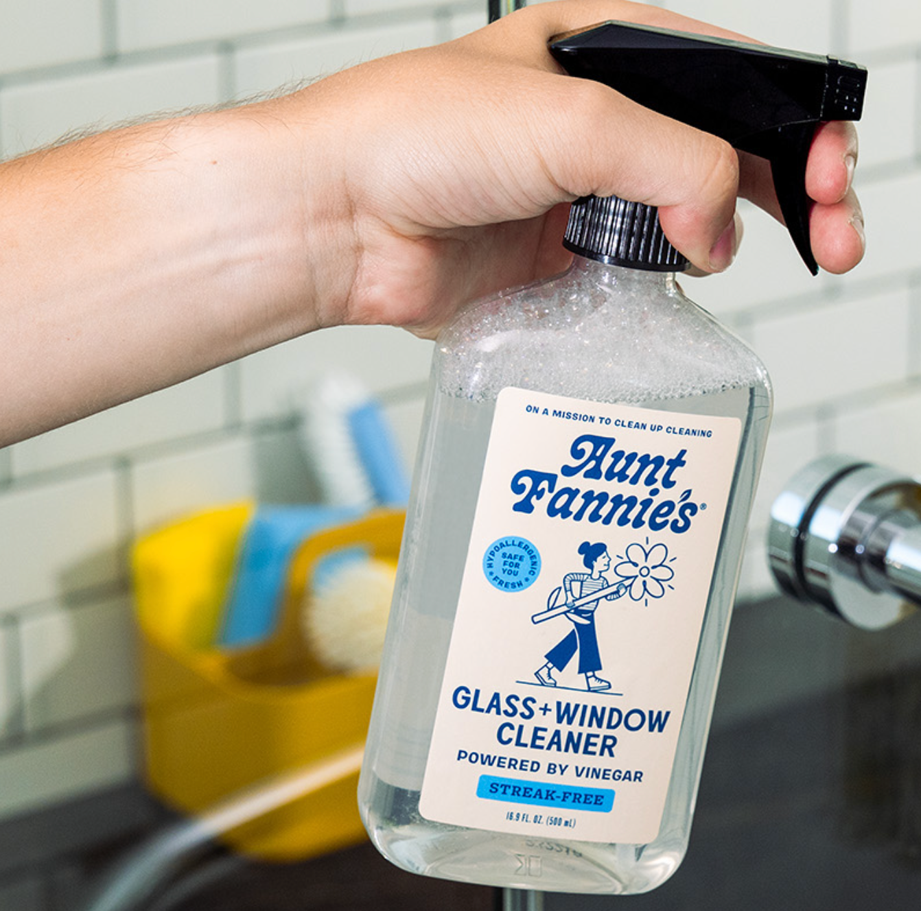 A hand holding a bottle of Aunt Fannie's Glass & Window Cleaner, powered by vinegar, with a label that features a cartoon character and text.