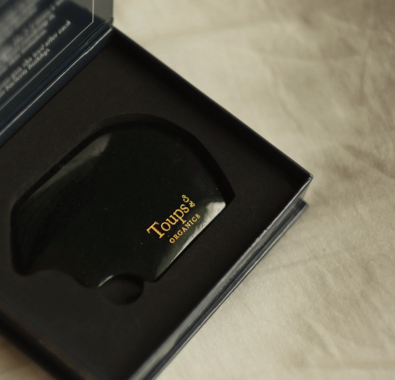 Hand holding a black curved hairbrush labeled "Toups," against a neutral beige background. The image conveys a minimalist and calm tone.
