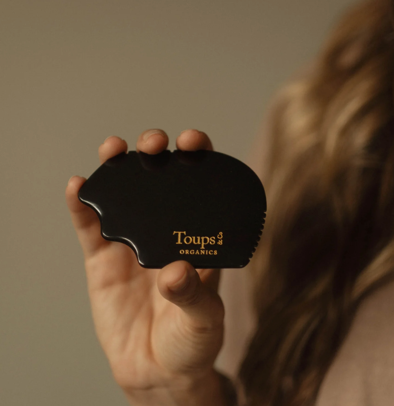 Hand holding a black curved hairbrush labeled "Toups," against a neutral beige background. The image conveys a minimalist and calm tone.