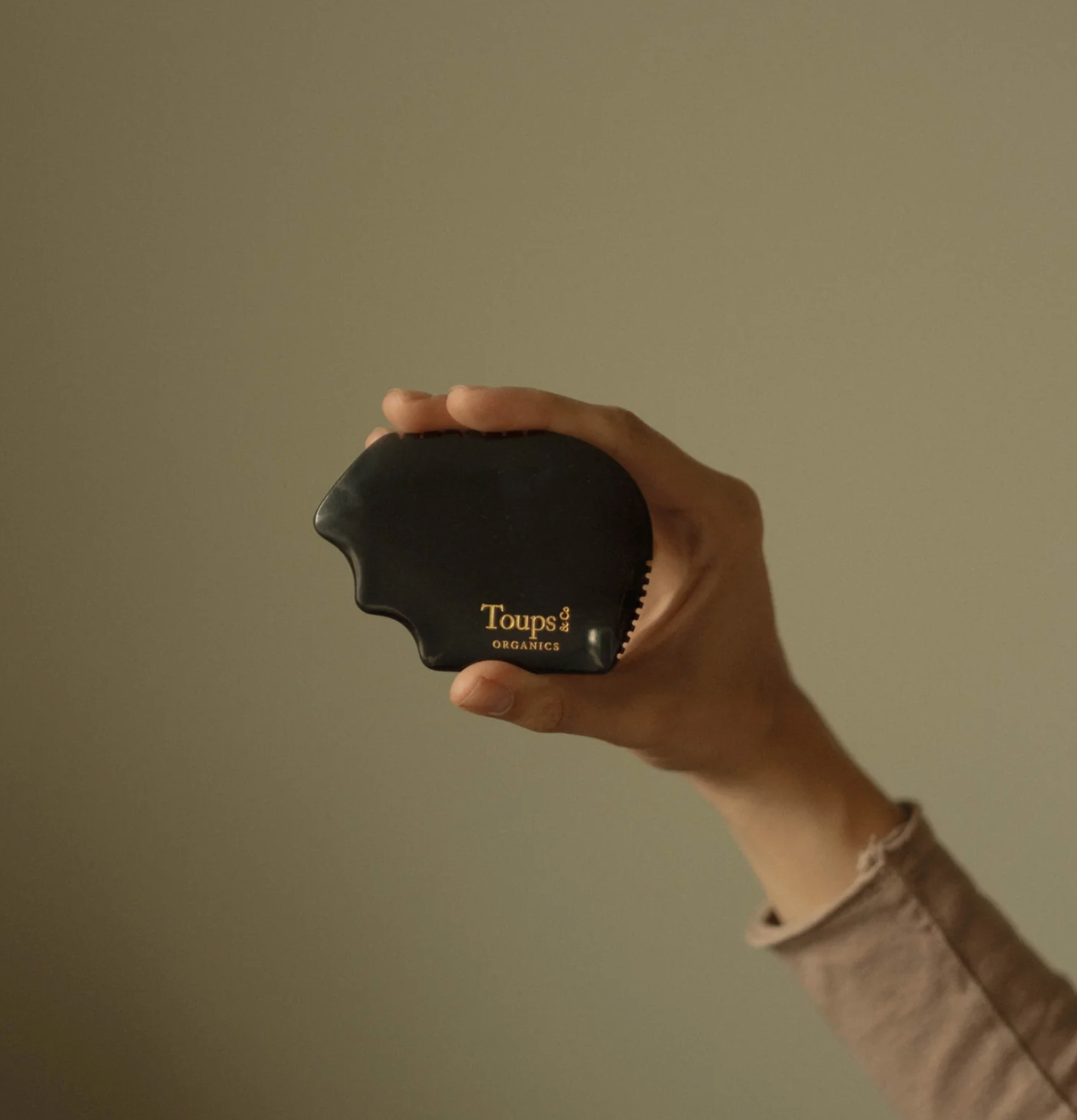Hand holding a black curved hairbrush labeled "Toups," against a neutral beige background. The image conveys a minimalist and calm tone.