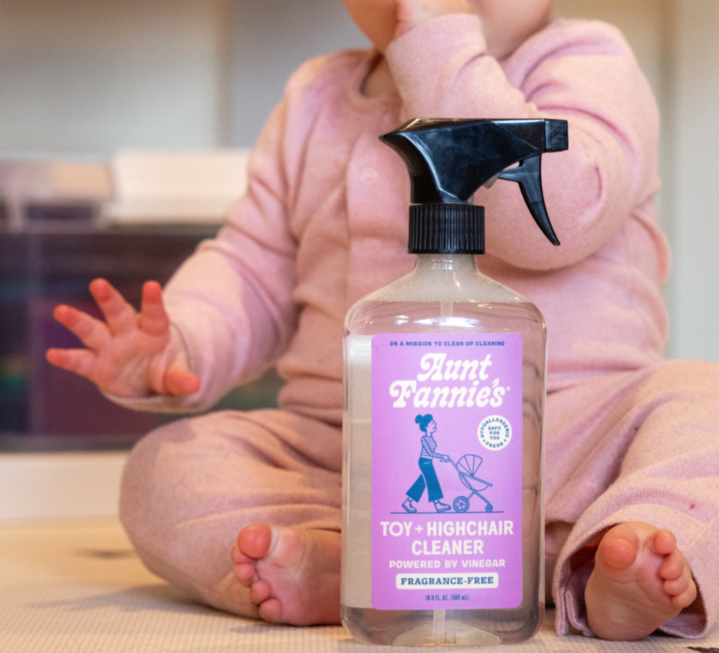 A clear spray bottle labeled 'Aunt Fannie's Toy + Highchair Cleaner' with a purple and green label, in front of a baby sitting on the floor.