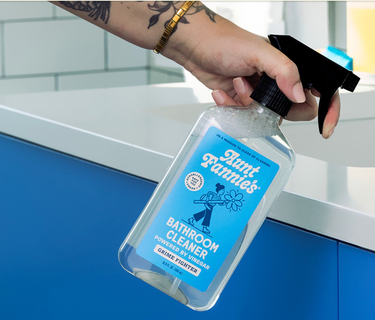 A hand holding a clear spray bottle labeled 'Bathroom Cleaner' with a blue and white design.