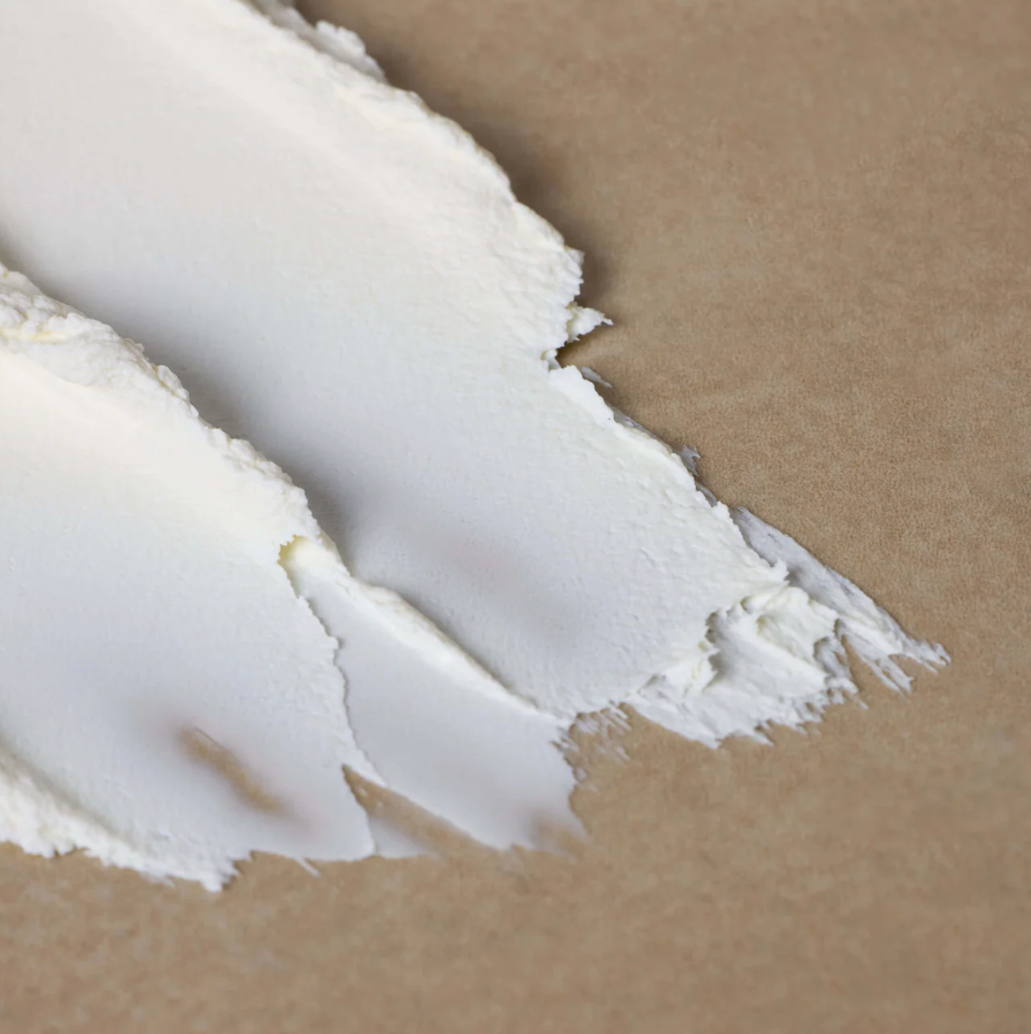 Close-up of two smooth, white cream smears on a beige surface. The creamy texture suggests softness and richness, creating a calming, neutral tone.