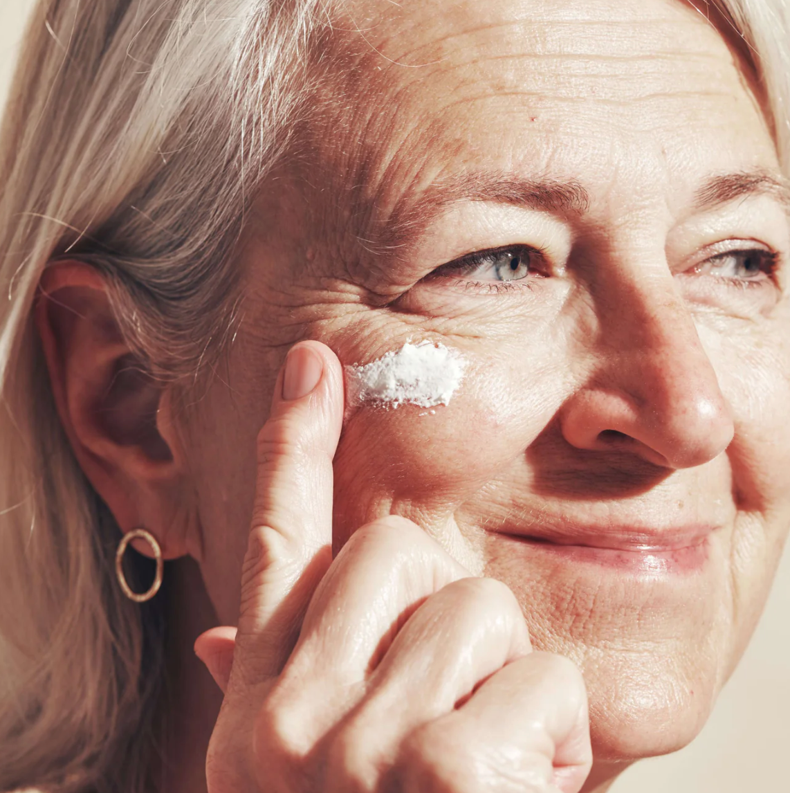 An older woman with grey hair is shown applying a white sun balm to her face, focusing on her eye area.