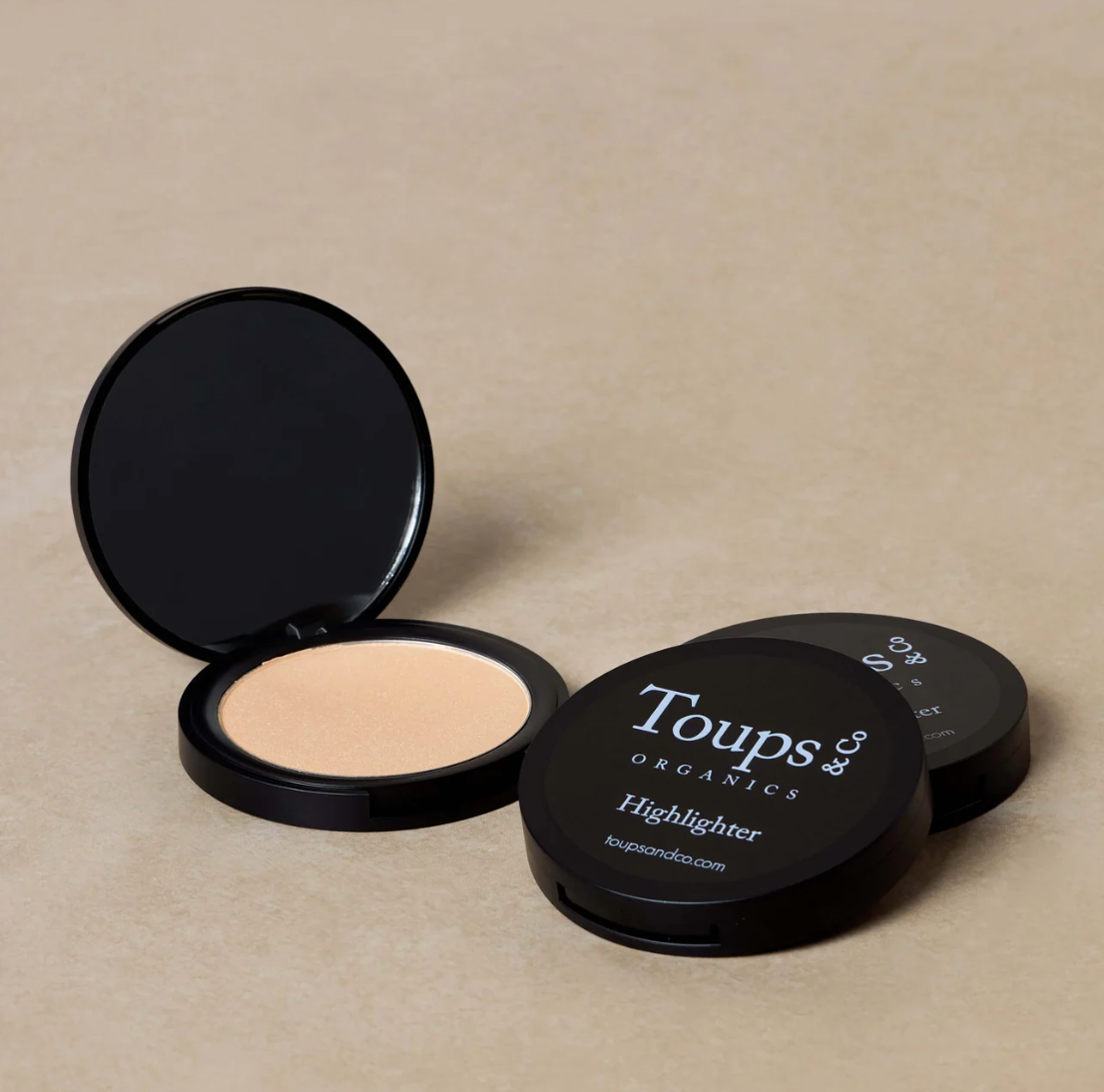 A compact highlighter by Toups & Co. is displayed open with the highlighter powder visible inside.
