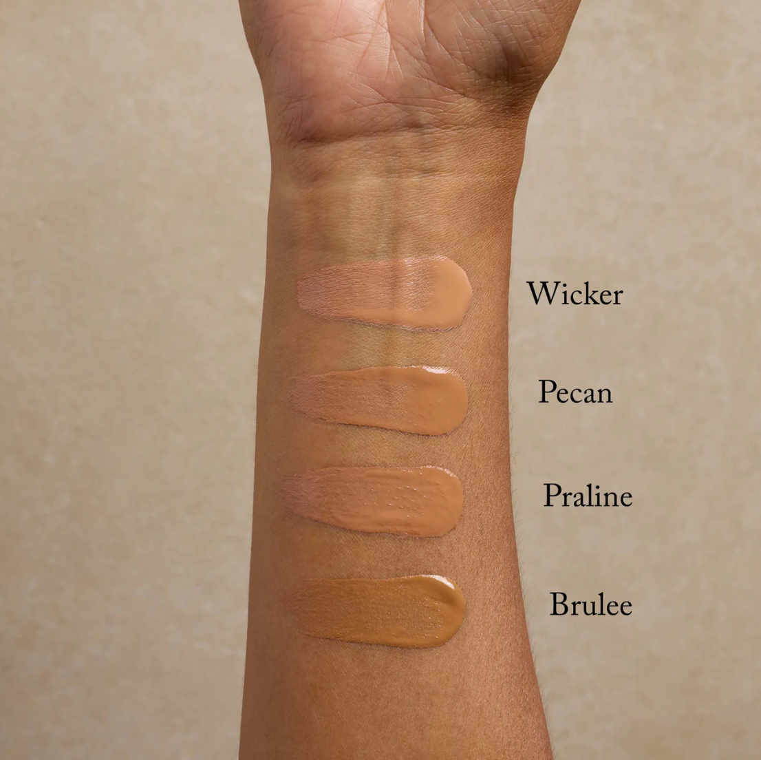 An arm with three different shades of liquid foundation applied in a strip, labeled Wicker, Pecan, and Brulee.