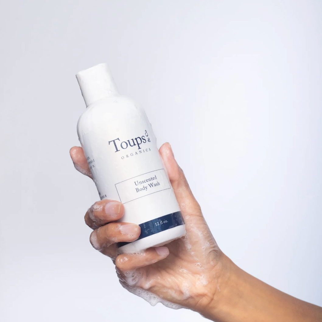 Rinsed hand holding a bottle of Toups Organics Unscented Body Wash against a light gray background