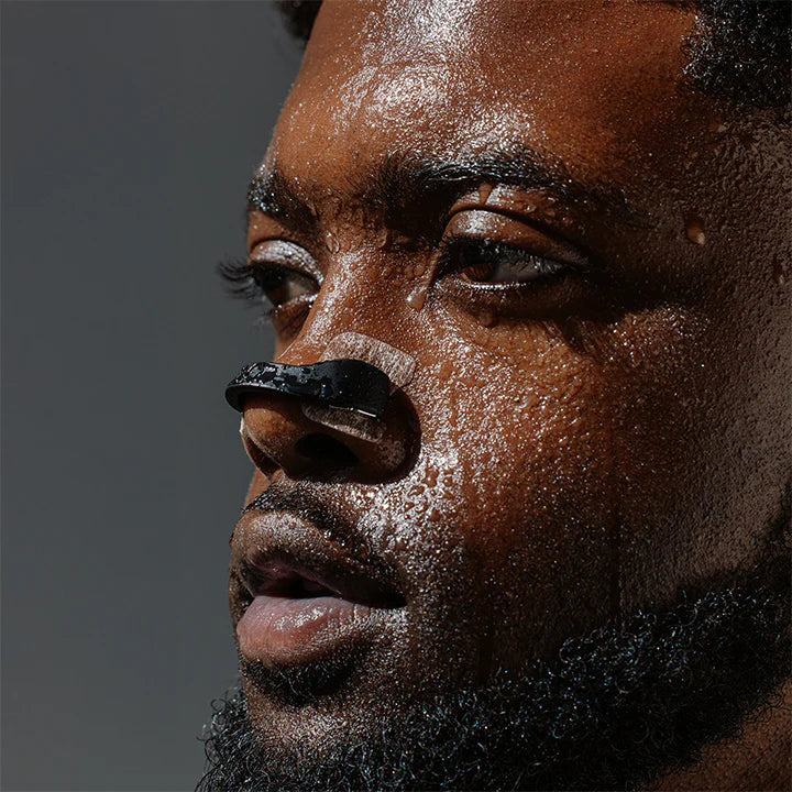 Close-up of a man's face, covered in sweat, wearing a black nose strip across the bridge of his nose, with a neutral expression.