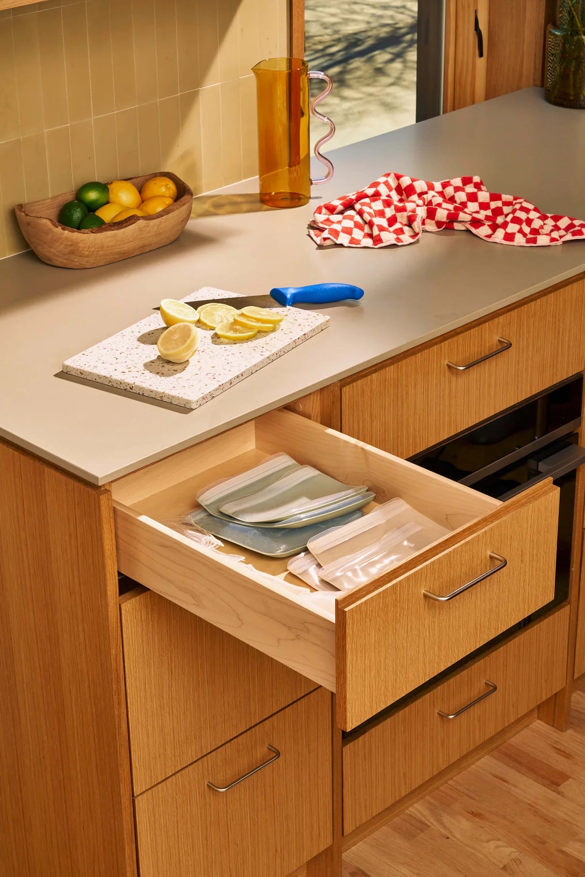 Kitchen with wooden cabinets, open drawer with utensils, lemons on cutting board, brown fruit bowl, red-checkered cloth, and a sunny ambiance.