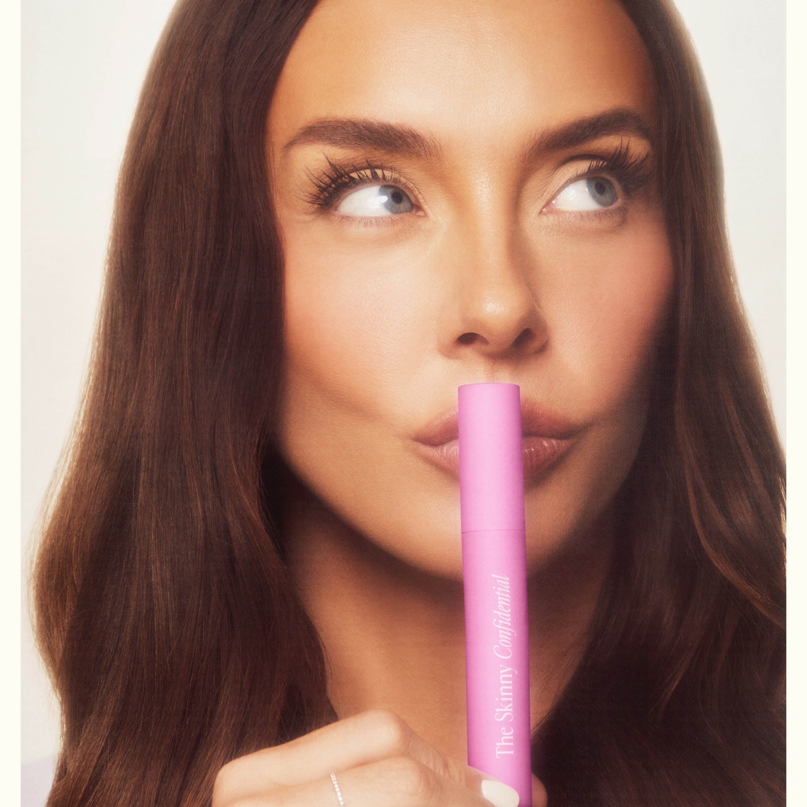 Woman with long brown hair holds a pink cosmetic tube near her lips. She gazes upwards thoughtfully. The mood is contemplative and serene.